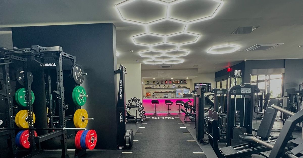 The bright gym floor at Solis Body Club: a Primal rack with colour plates, machines under the hexagon lights and the smoothie bar at the back